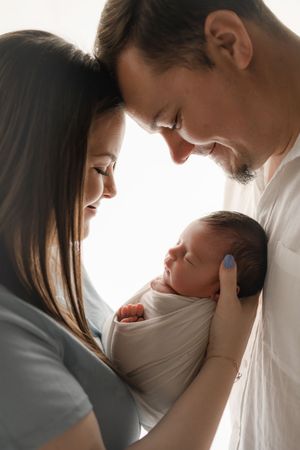 Smiling parents holding their swaddled newborn baby together in soft white light.