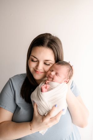 Brunette mother in blue dress smiling warmly while holding swaddled sleeping newborn baby.