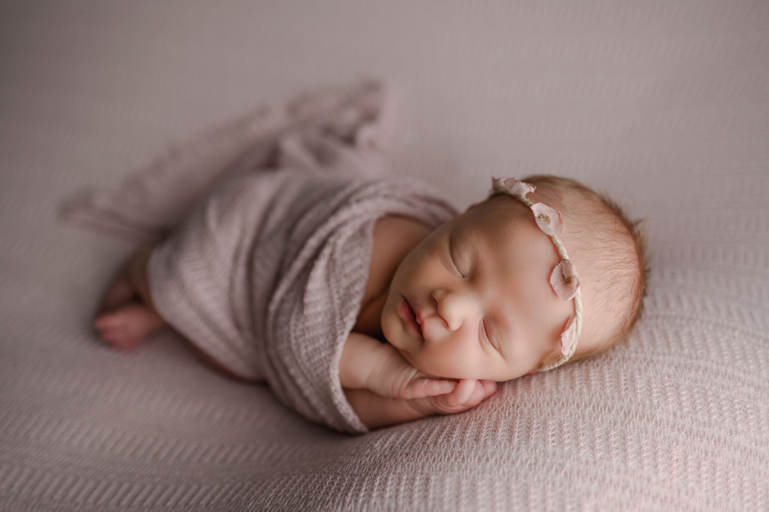 Sleeping newborn baby wrapped in lavender swaddle with matching headband on soft gray backdrop.