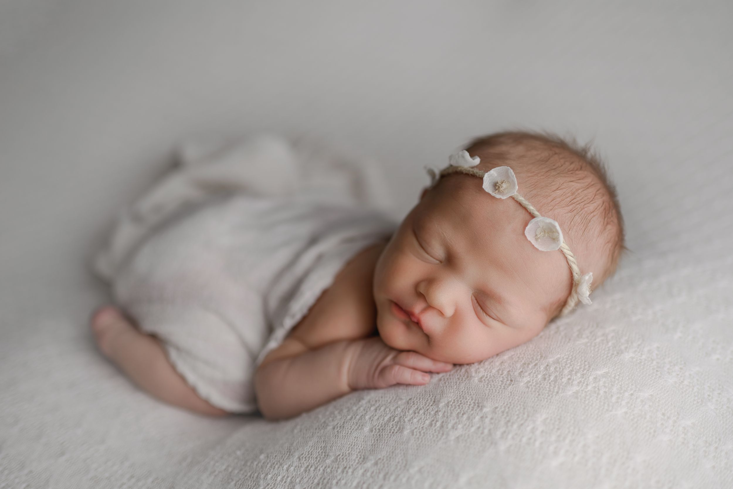 Peacefully sleeping newborn baby in white wrap with delicate floral headband on soft white blanket.