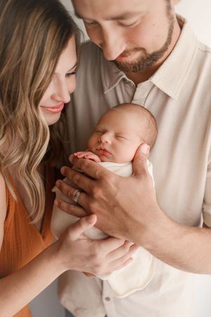 Loving parents lean in close together cradling their sleeping swaddled newborn baby.