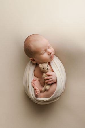 Sleeping newborn baby swaddled in cream wrap cuddles a tiny knit teddy bear toy.