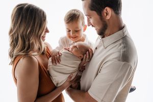 Happy parents with toddler son admire their swaddled newborn baby in a bright studio.