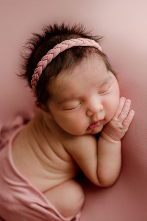 Sleeping newborn baby girl in pink wrap and braided headband on pink background.