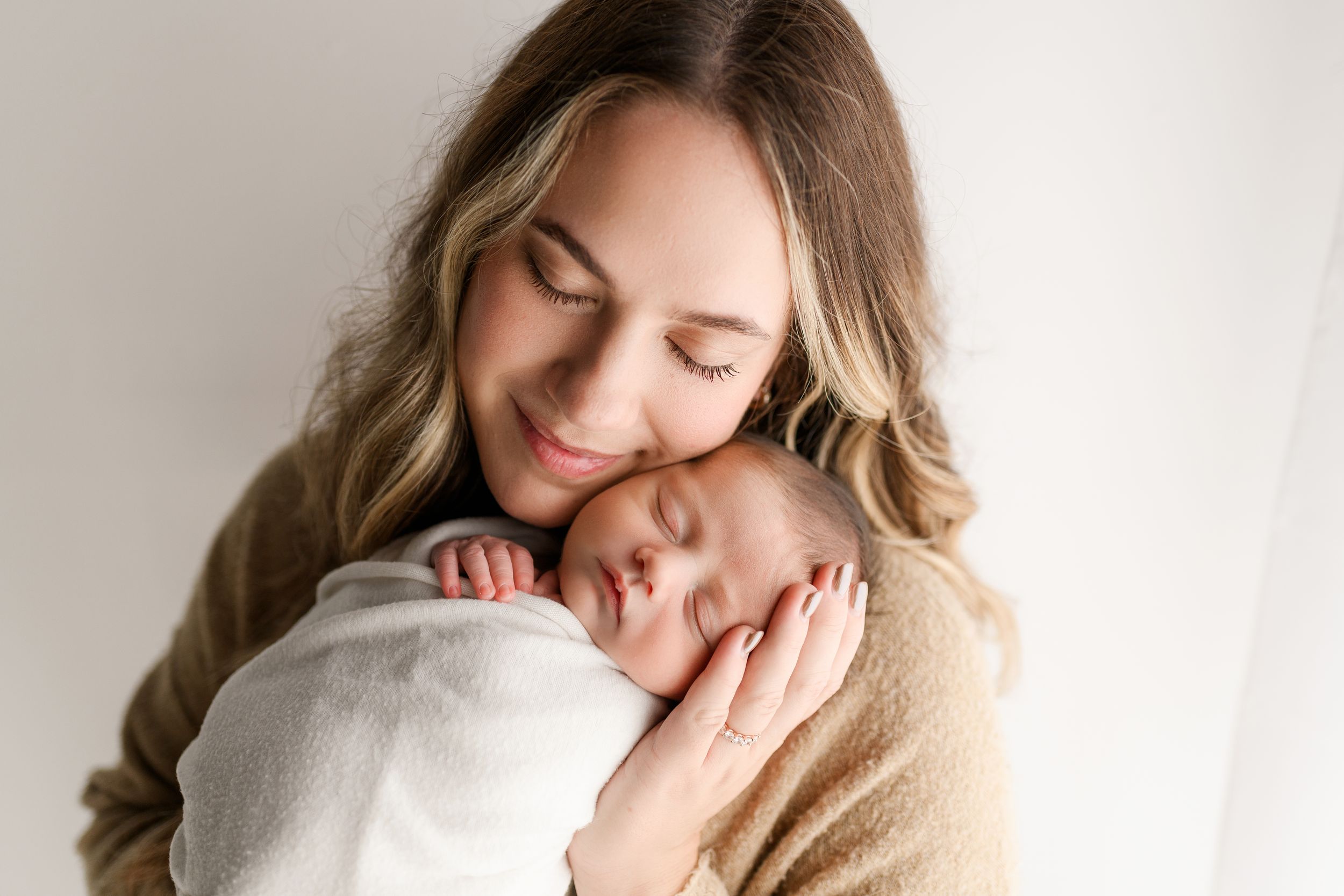Mother tenderly holds sleeping newborn baby wrapped in white, eyes closed, smiling softly in a bright studio.