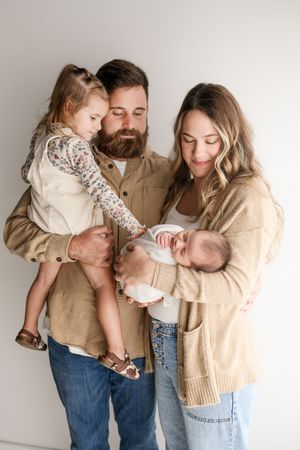 Family portrait with bearded father holding toddler girl and mother cradling swaddled newborn in warm neutral tones.