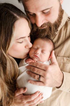 Parents lovingly kiss and cuddle their swaddled sleeping newborn baby together in a tender portrait.