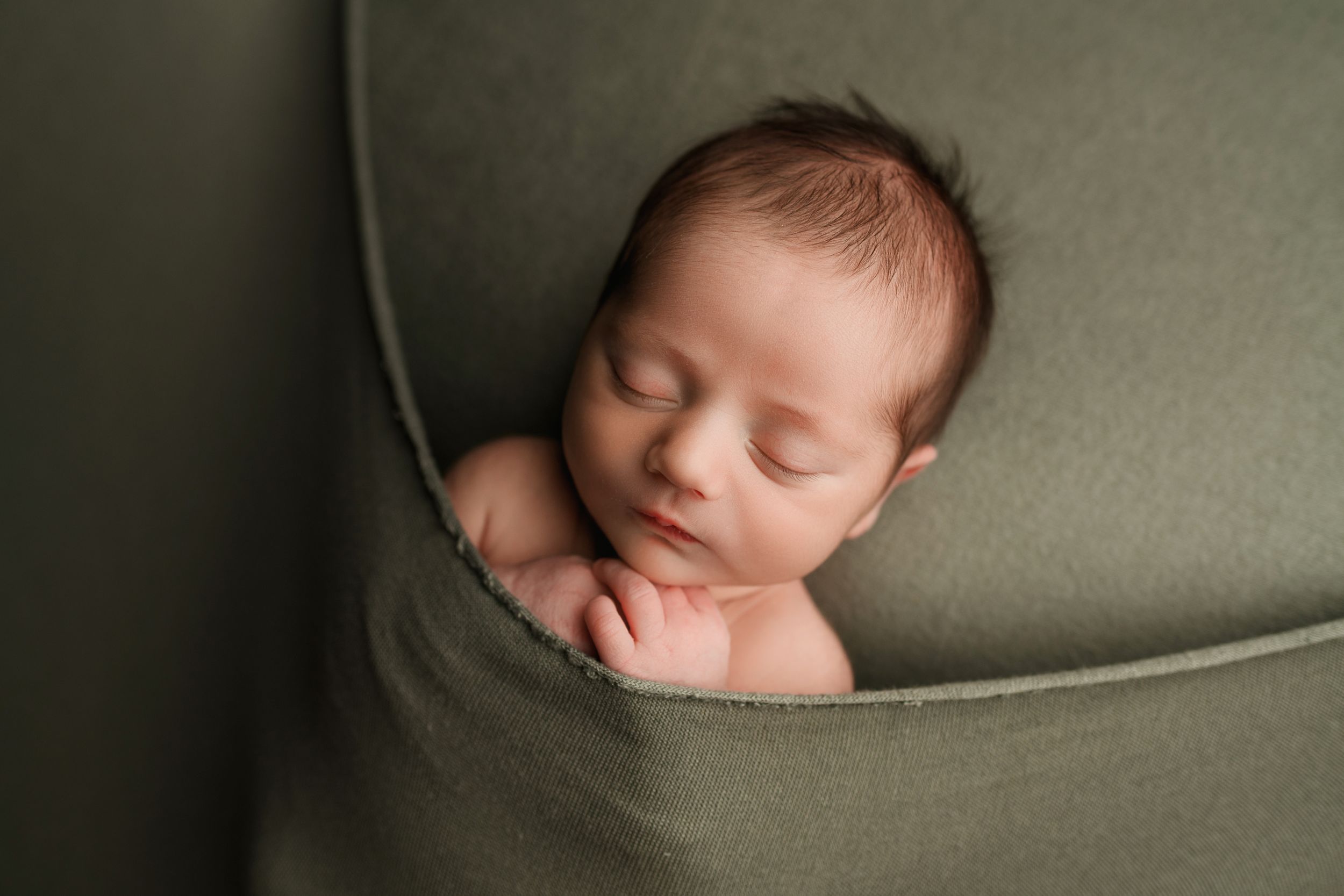 Peaceful sleeping newborn baby nestled in olive green fabric wrap in a cozy bowl pose.