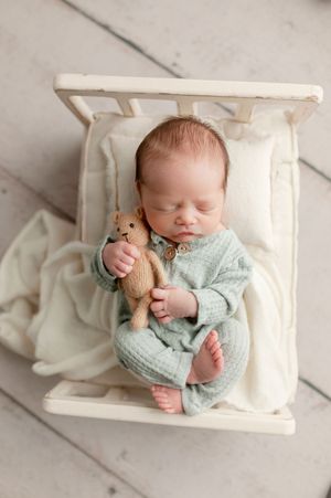 Sleeping newborn in mint green knit outfit holding teddy bear in a miniature white wooden bed.