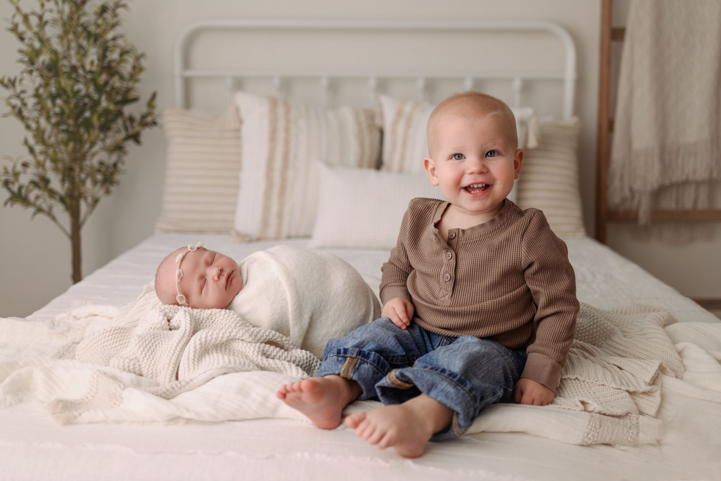 Smiling toddler boy sits beside sleeping swaddled newborn sibling on white bed.
