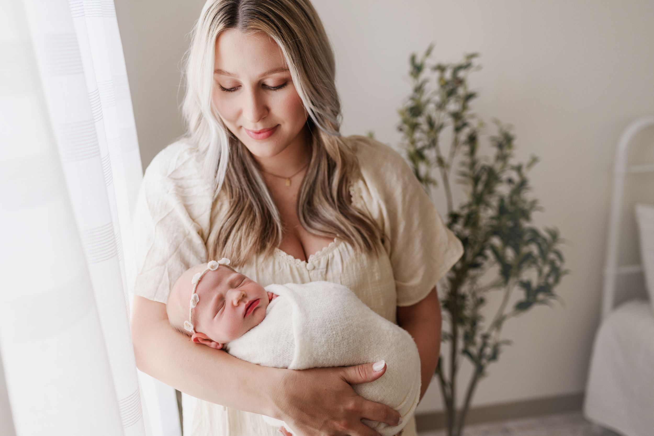 Blonde mother in cream dress tenderly holds sleeping swaddled newborn baby.