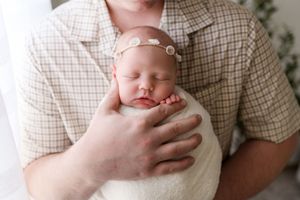 Father in plaid shirt gently cradles sleeping swaddled newborn with floral headband.
