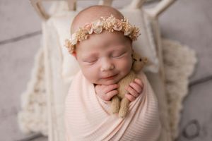 Smiling sleeping newborn in pink wrap and floral crown holds knit teddy bear.