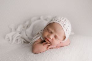 Peaceful sleeping newborn baby in white knit bonnet posed on soft white blanket.