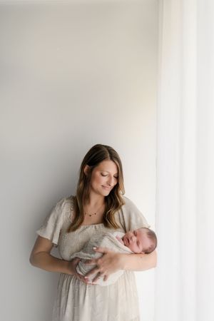 Smiling mother in beige dress holding newborn baby near bright white curtained window.