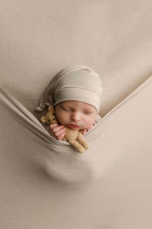 Peaceful newborn baby in beige knit hat sleeps in a fabric hammock holding a teddy bear.