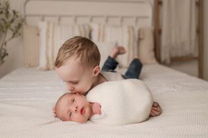 Older toddler brother lying on white bed kissing sleeping swaddled newborn sibling tenderly.