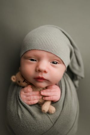 Alert newborn baby swaddled in sage green wrap wearing matching knit hat, holding tiny teddy bear.