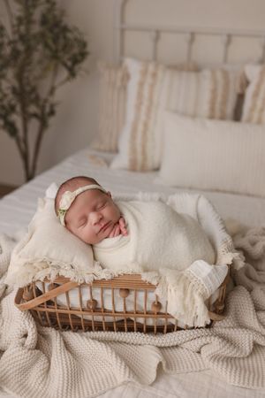 Sleeping newborn baby girl swaddled in cream wrap rests in a wicker basket on a bed.