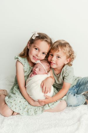 Two young siblings smile while gently holding their swaddled newborn baby sister.