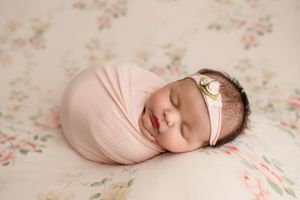 Smiling sleeping newborn girl swaddled in pink wrap with floral headband on floral backdrop.