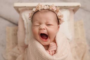 Yawning newborn baby wrapped in blush pink swaddle wearing floral crown in a tiny wooden bed.
