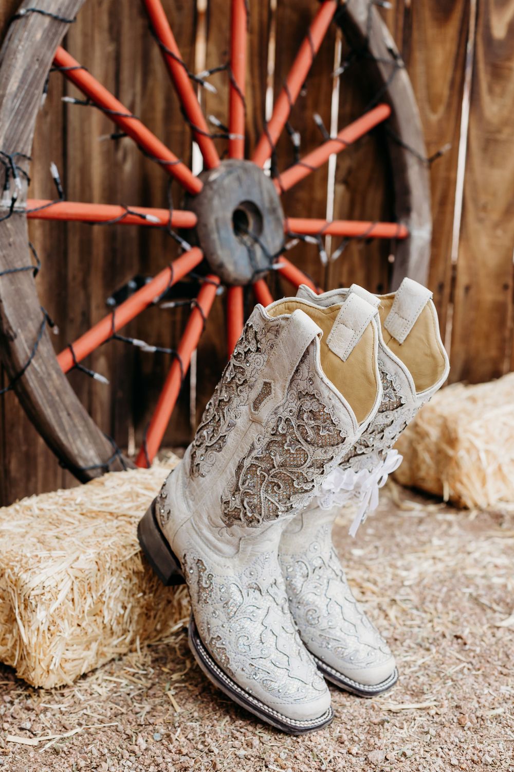 Ornate white cowboy boots with intricate embroidery displayed in front of a rustic wagon wheel.