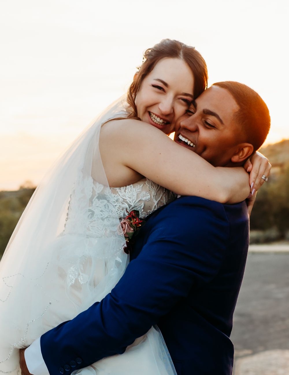 Joyful wedding couple embrace in warm sunset light with bride in white lace dress.