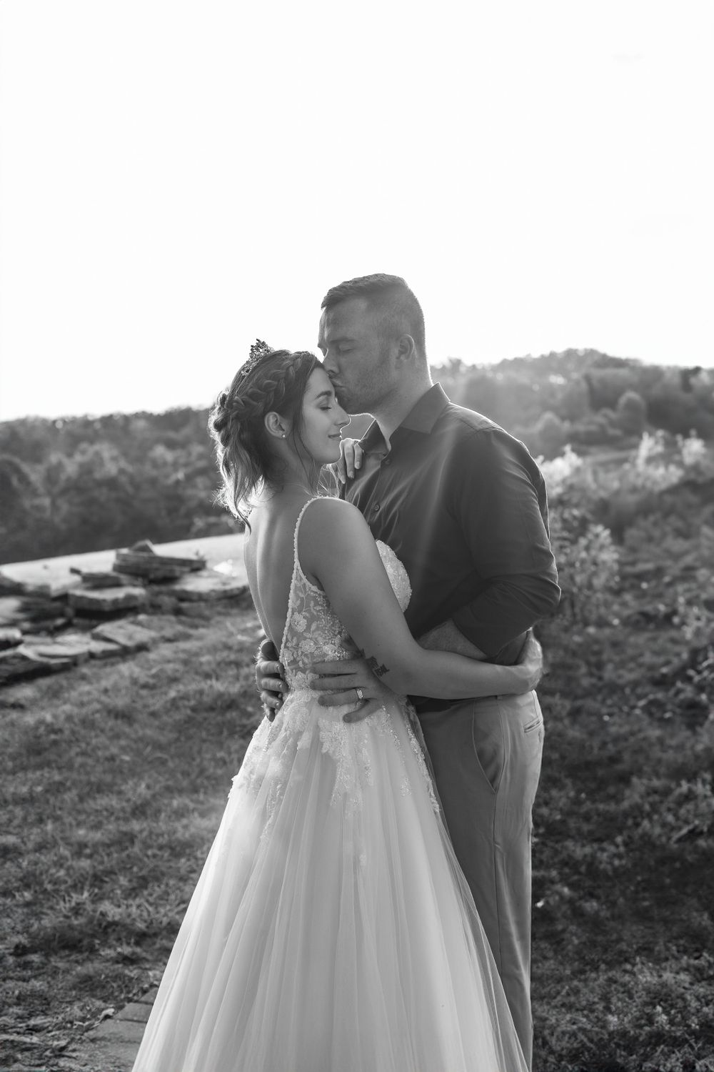A romantic black and white wedding photograph of a couple embracing outdoors at sunset on a hillside.
