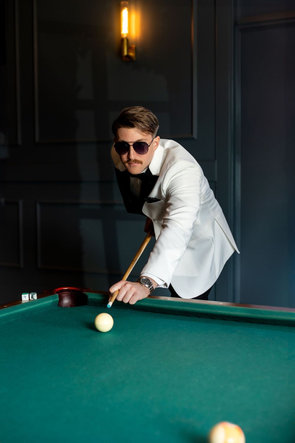 Stylish person in white blazer and sunglasses leans over pool table to take a shot in dimly lit room.