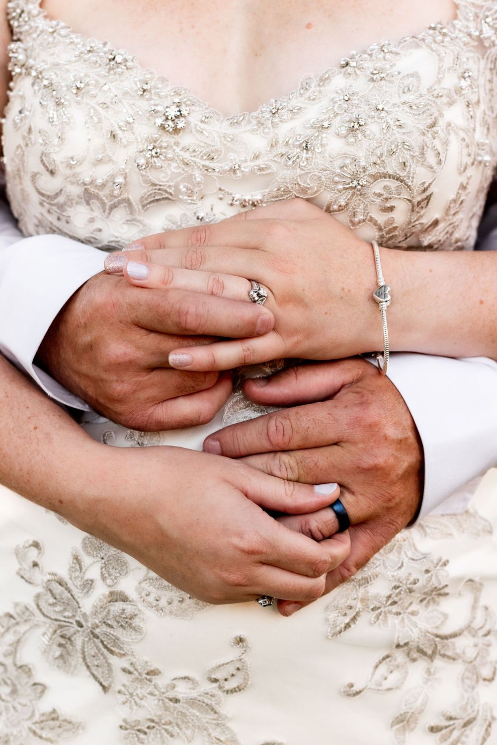Close up of wedding rings and hands against an ornate beaded white wedding dress.