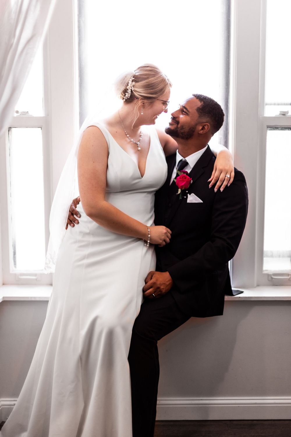 A couple in wedding attire share an intimate moment by a window, with the bride in white and groom in black suit.