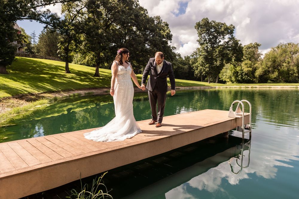 A couple walks along a wooden dock by a serene lake surrounded by lush greenery on a sunny day.