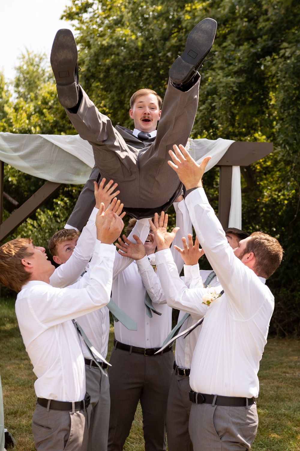 Groomsmen in white shirts and gray vests playfully toss and catch someone during an outdoor wedding celebration.