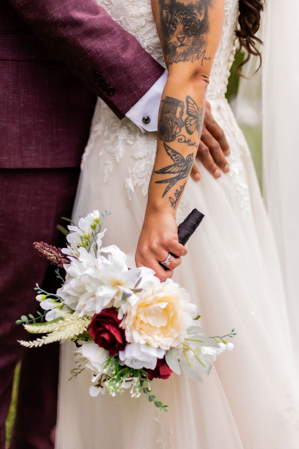 A close-up of tattooed arms holding a wedding bouquet with white and burgundy flowers against a white dress.