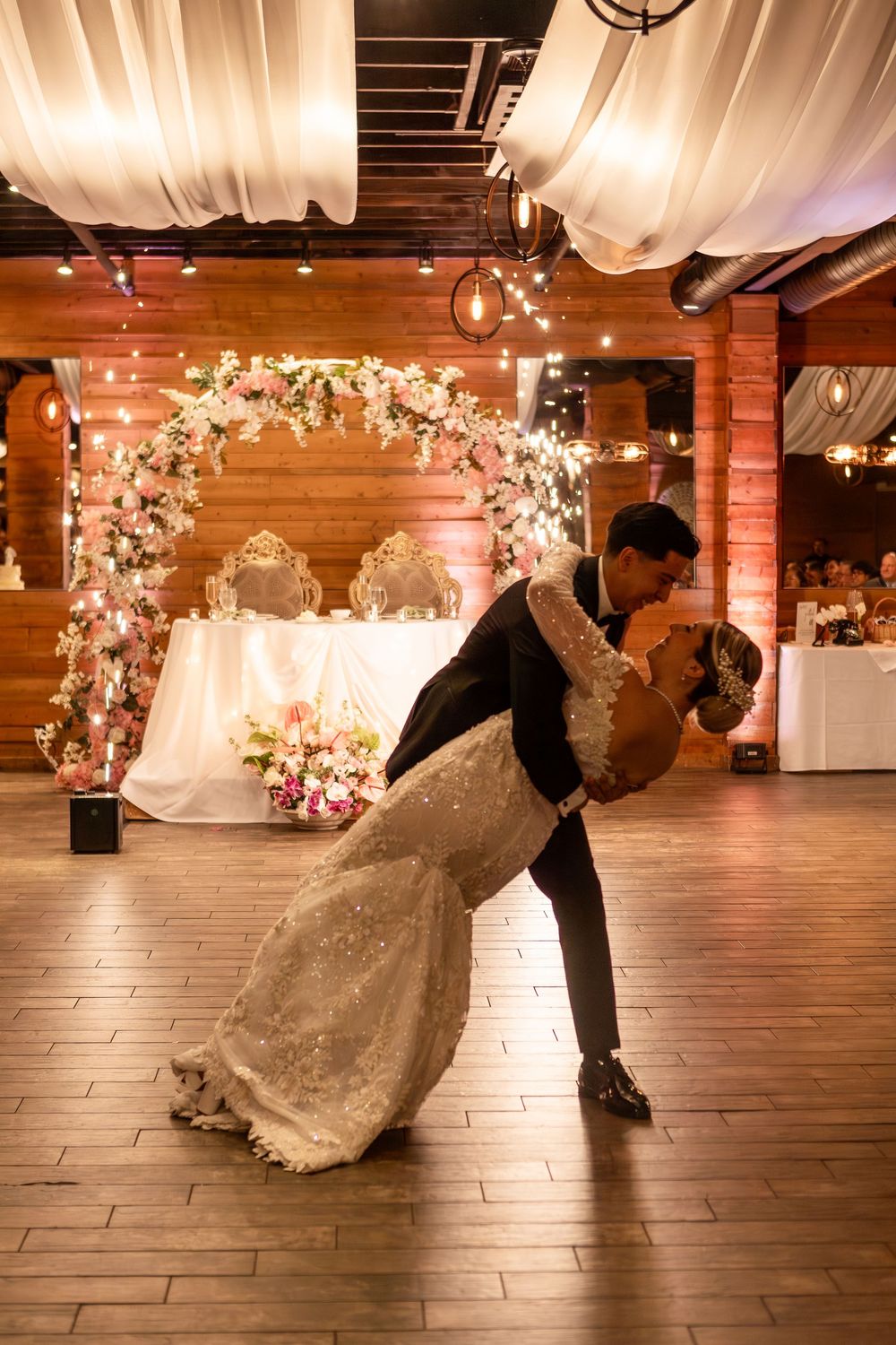 A couple performs a romantic dip dance move in a rustic venue decorated with floral arches and white draping.