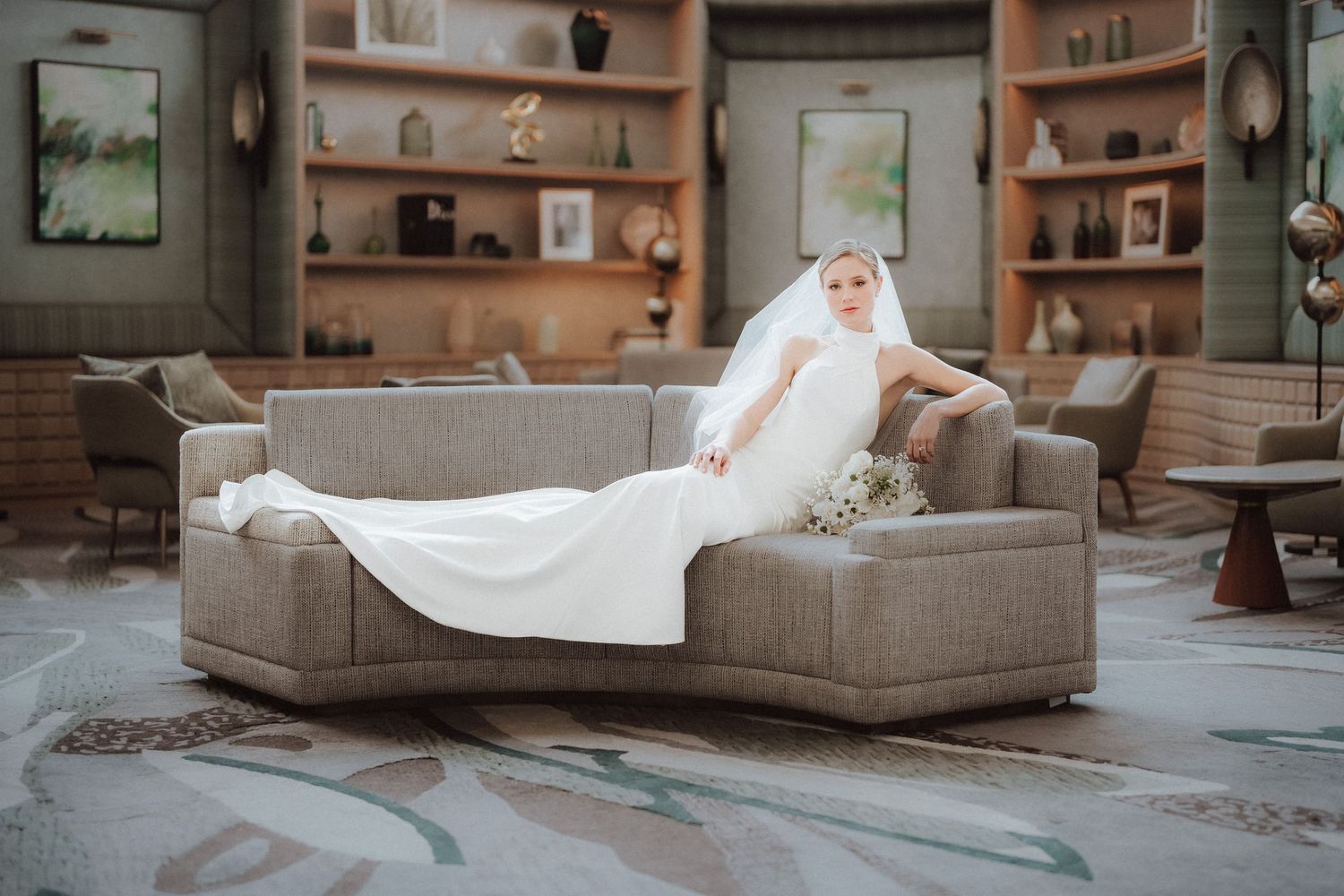A white cat lounges on a gray sectional sofa in a cozy living room with wooden shelves in the background.