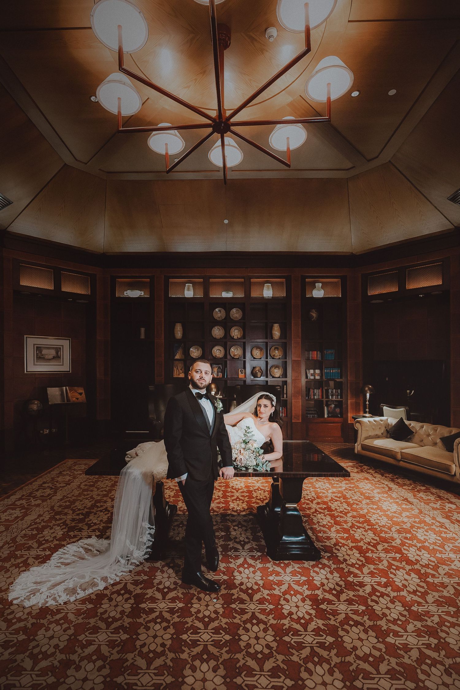 A wedding couple poses in a luxurious wood-paneled room with ornate red carpeting and dramatic overhead lighting.