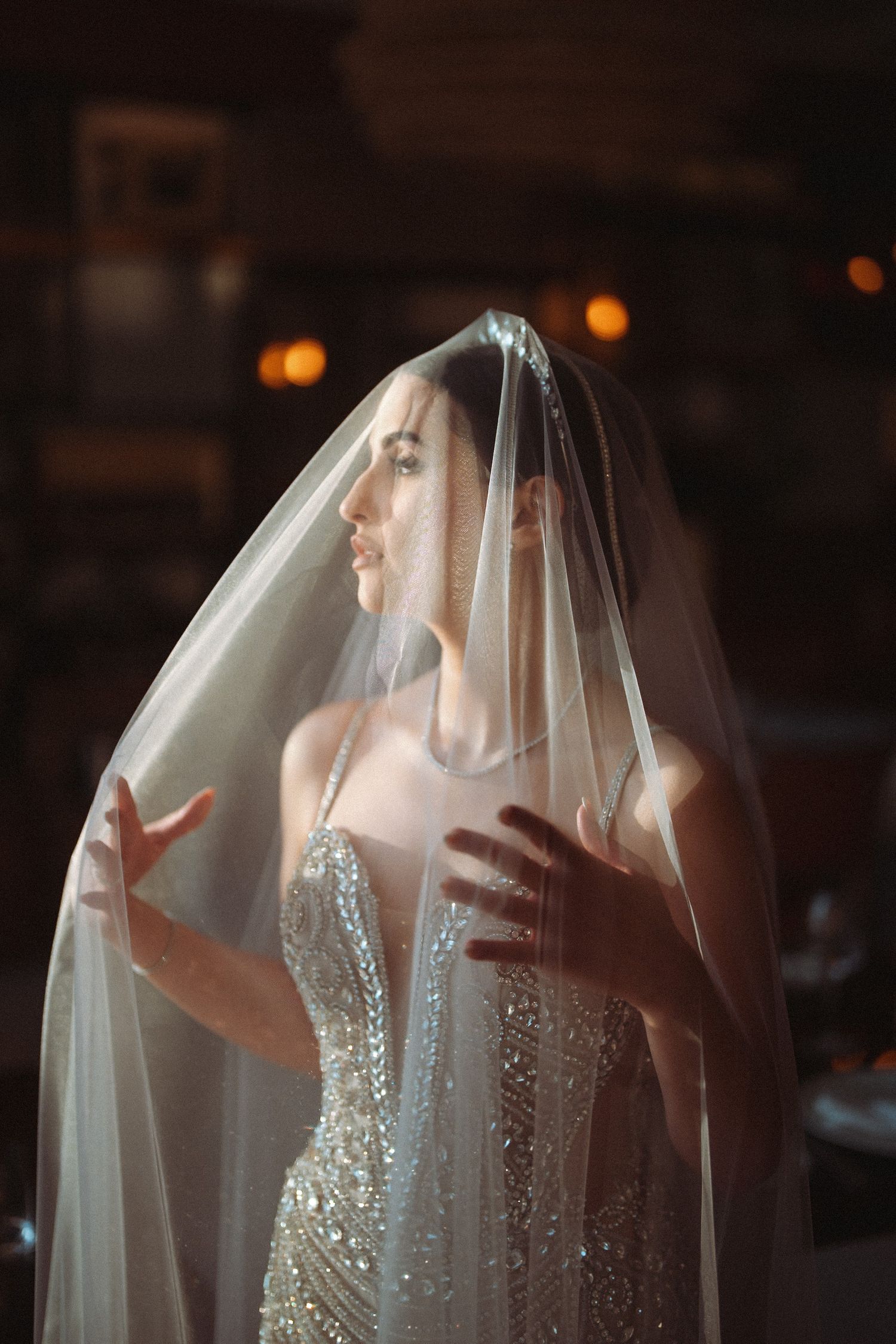 A bride in a white veil poses behind sheer fabric creating an artistic silhouette effect in a dark room.