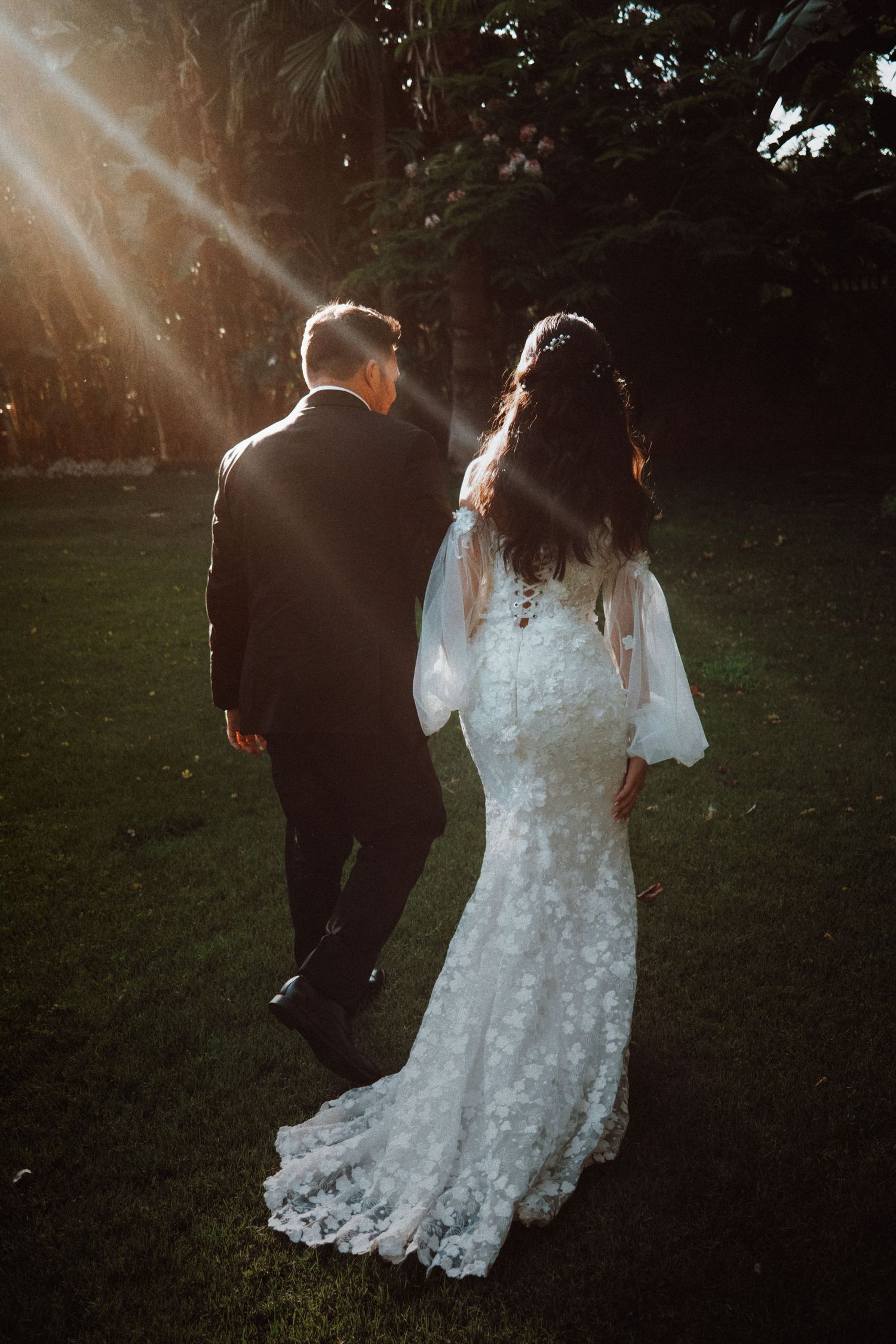 A couple in wedding attire walks through a sunlit garden with romantic lens flare and dreamy atmosphere.