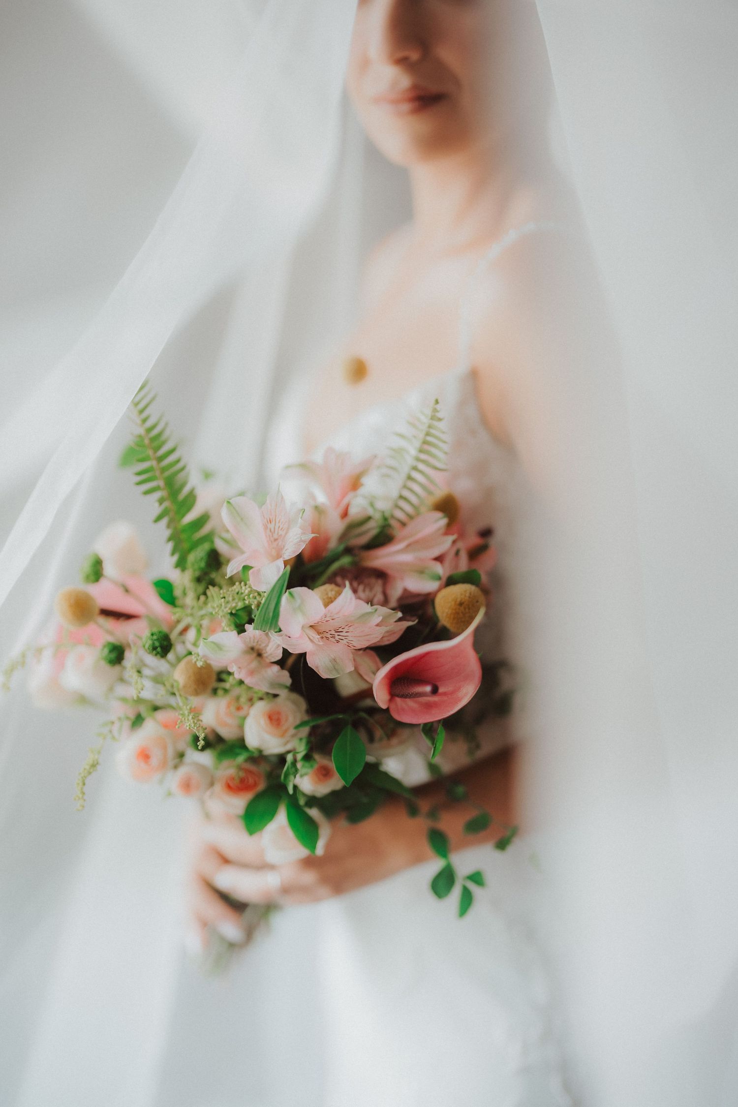 A dreamy bridal portrait shows a soft-focus bouquet of pink calla lilies, roses and ferns through a delicate white veil.