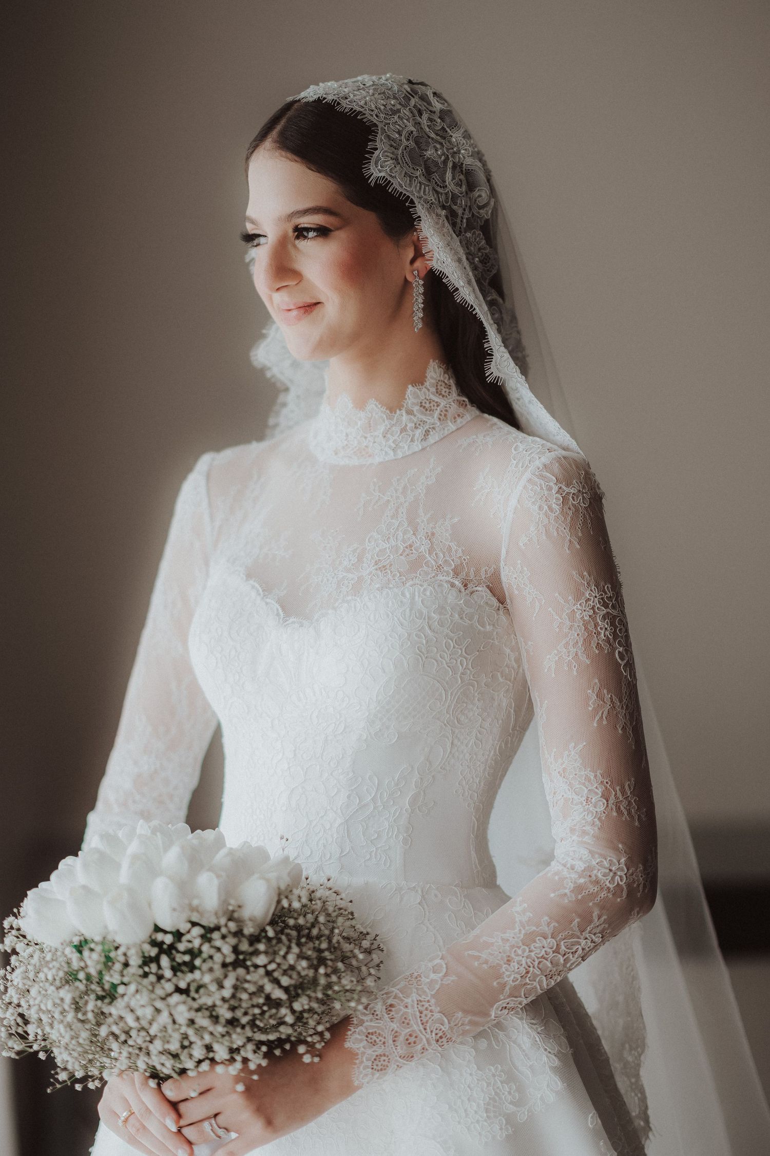 A bride in a classic white lace long-sleeve wedding gown holds a delicate baby's breath bouquet in soft natural lighting.