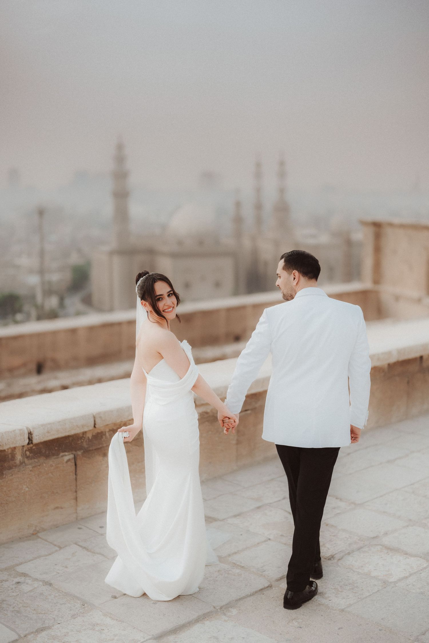 A couple in wedding attire dances gracefully on a rooftop terrace with a misty cityscape in the background.