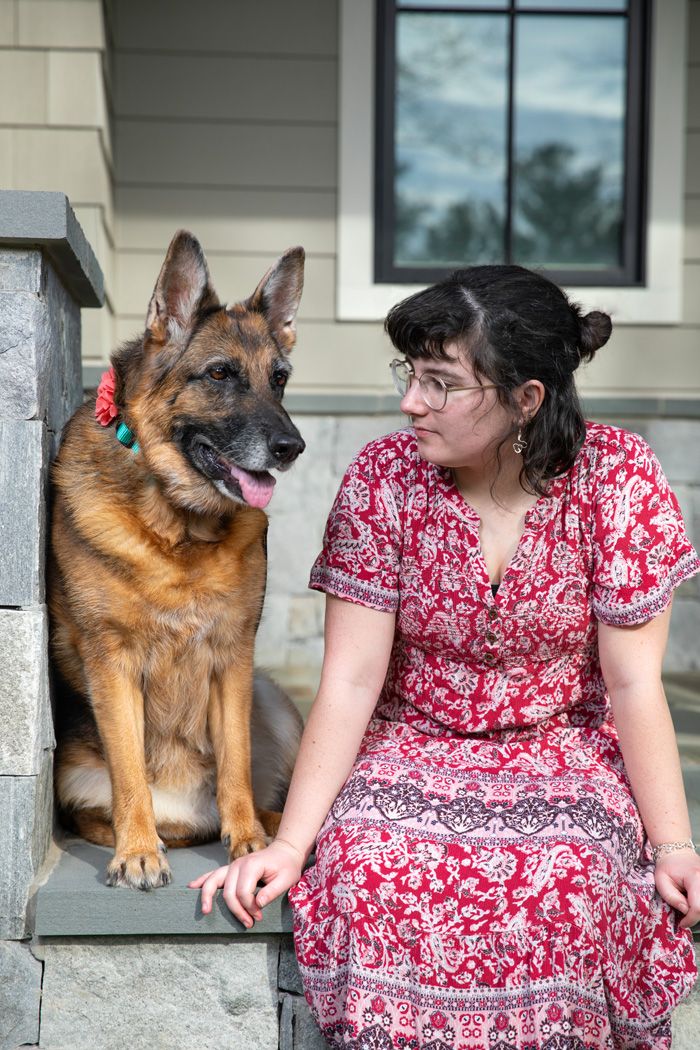 A German Shepherd dog sits beside someone in a red patterned dress on outdoor steps.