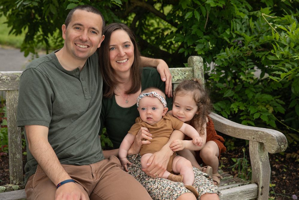 A family sits together on a wooden garden bench surrounded by greenery.