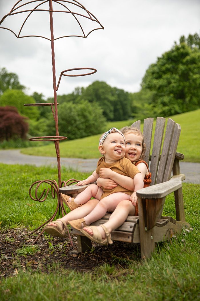 Two children share laughter while sitting in an Adirondack chair under a decorative metal umbrella.