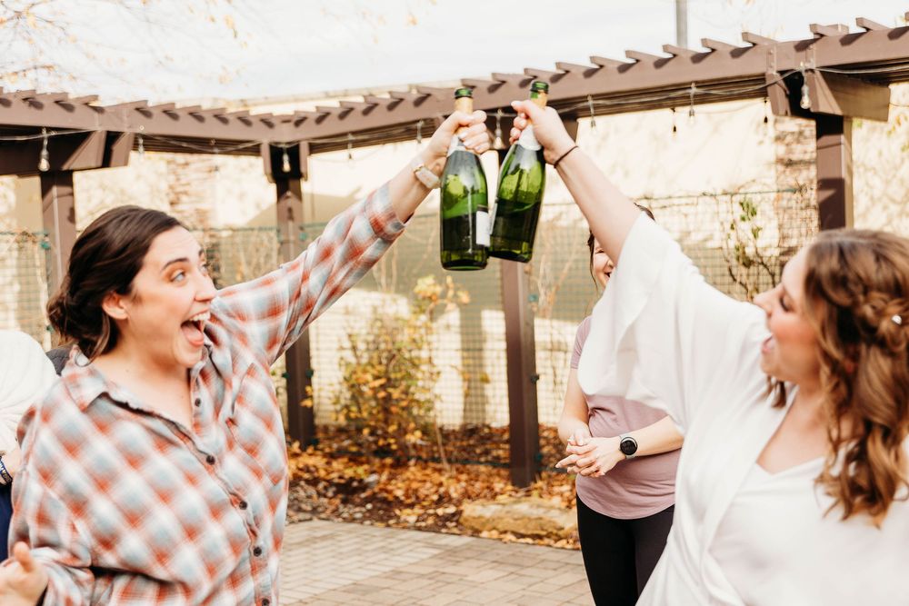 Two friends laugh and cheer while clinking beer bottles together under a wooden pergola in an outdoor setting.