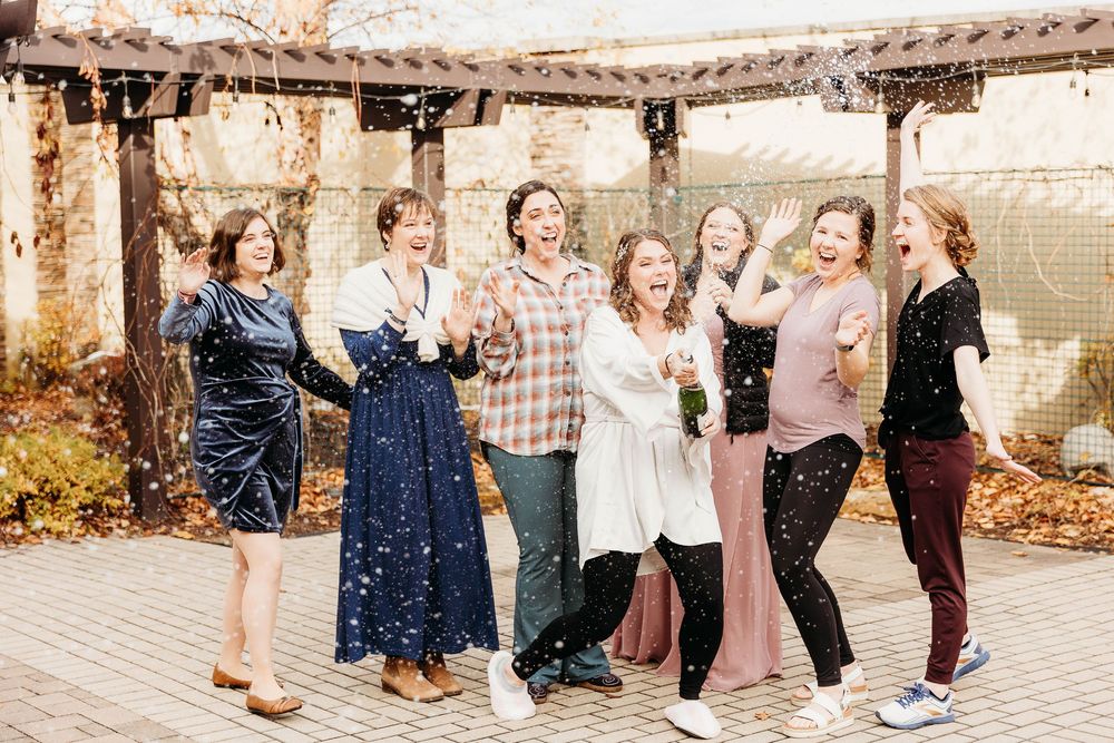 A joyful group celebrating together outdoors under a pergola wearing casual fall attire in mixed colors.