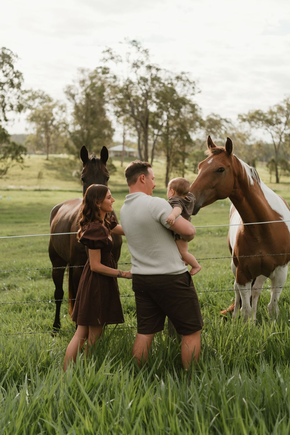 A family with a baby stands between two horses in a lush green paddock, sharing a tender moment outdoors.