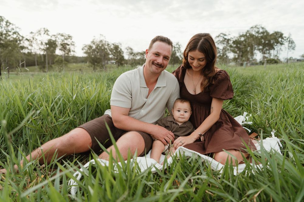 A happy family of three sits in a lush green field, parents smiling with their baby on a sunny day.
